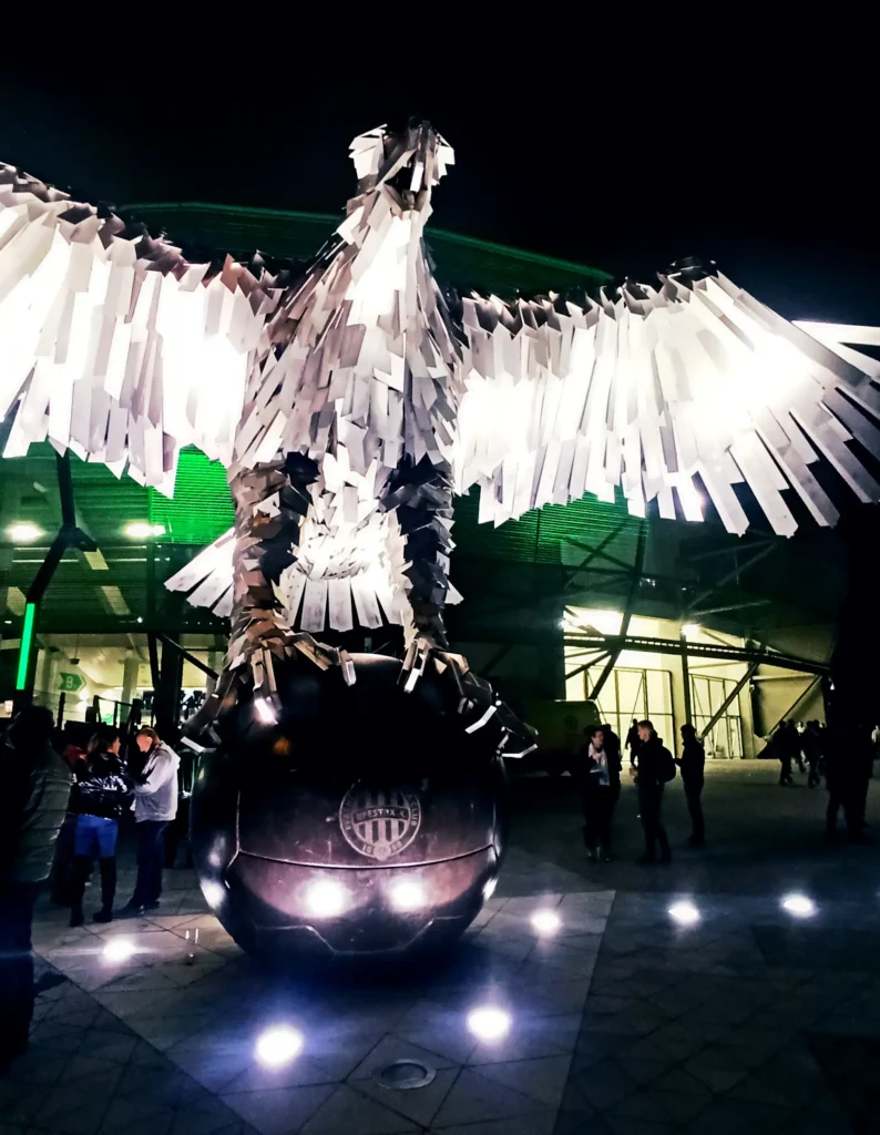Fradi-Sas Escultura monumental iluminada de acero inoxidable del "Fradi Sas" (Águila del Fradi) posada sobre un balón de fútbol frente a la entrada del estadio Groupama Arena por la noche.
