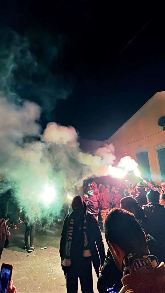 Green-Monsters-Bengalas Corteo nocturno de los ultras Green Monsters en Budapest iluminado por bengalas verdes y denso humo blanco, con aficionados vistiendo ropa oscura y bufandas.