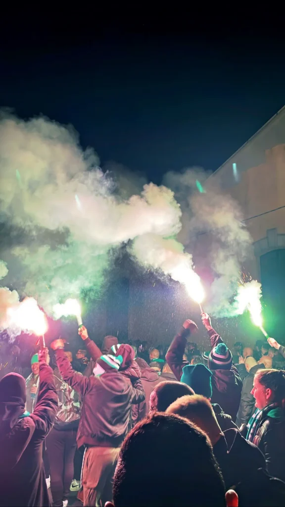 Green-Monsters-Pirotecnia Ultras de los Green Monsters sosteniendo bengalas verdes y rojas envueltos en humo denso durante una previa nocturna en Budapest.