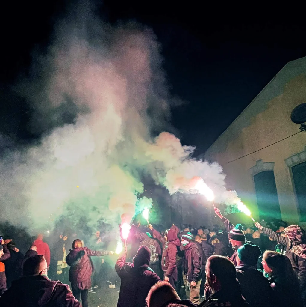 Green-Monsters-Pyro Plano abierto de ultras del Ferencváros alzando bengalas verdes y rojas junto a un edificio amarillo, envueltos en una densa nube de humo nocturno.