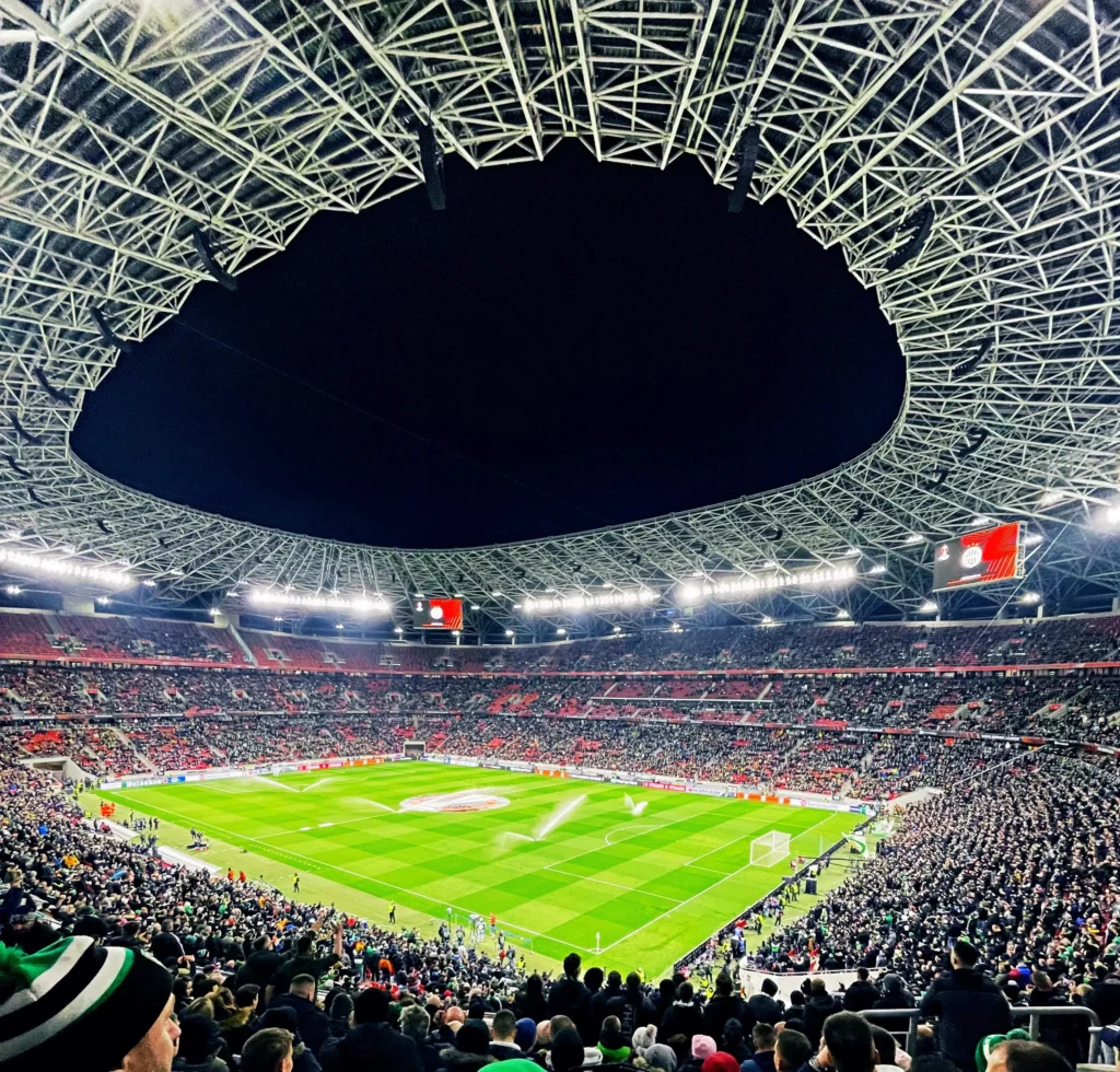 Puskas-Arena Vista panorámica nocturna desde la grada del Puskás Aréna repleto de aficionados, destacando la compleja estructura del techo y el terreno de juego iluminado.