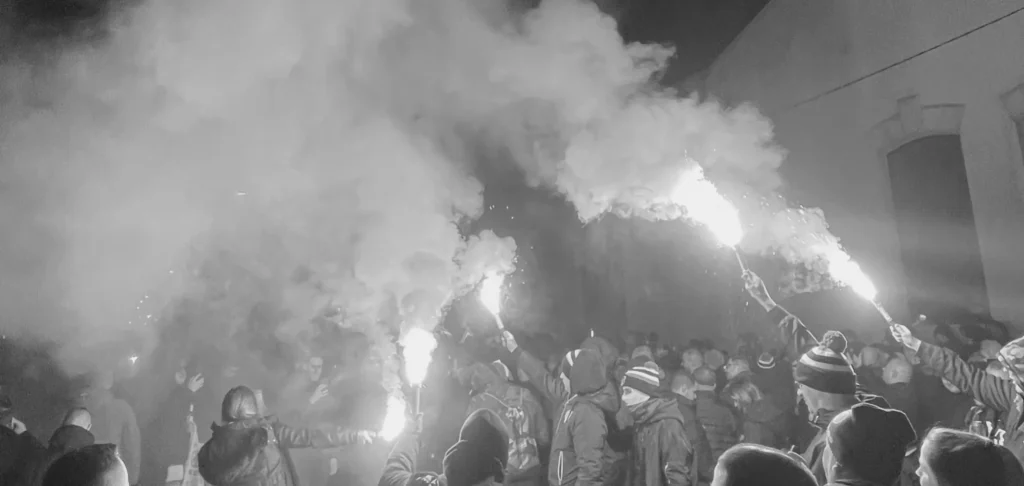 Fotografía artística en blanco y negro de ultras alzando bengalas, creando un fuerte contraste entre la luz del fuego, el humo denso y la oscuridad de la noche.