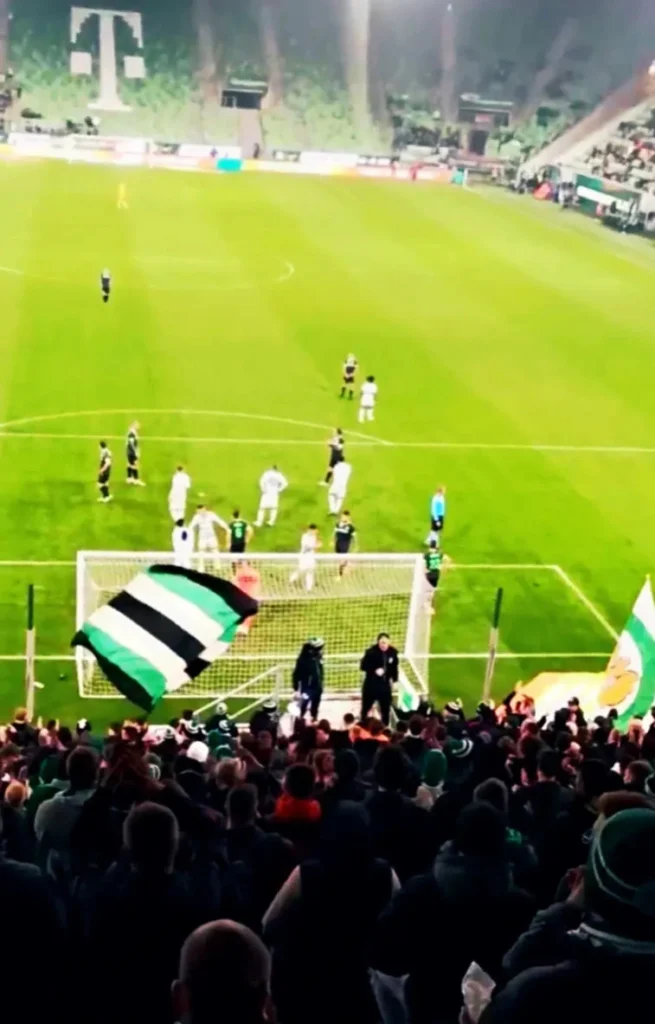 Sektor-B-Groupama-Arena Vista subjetiva desde el interior del Sektor B en el Groupama Arena, mostrando a los ultras de espaldas con banderas verdiblancas y el terreno de juego al fondo.