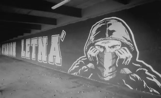 Fotografía en blanco y negro de un gran mural de graffiti en el estadio del Sparta Praga. Muestra a un hooligan encapuchado cubriéndose el rostro junto al texto "STADION LETNÁ" en escala de grises.