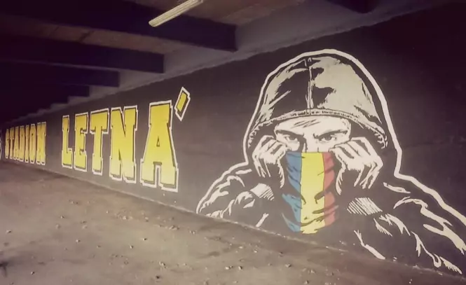 graffiti-hooligans-sparta-praga Mural de graffiti bajo las gradas del estadio que muestra a un hooligan del Sparta Praga encapuchado cubriéndose el rostro con una bufanda tricolor (azul, amarillo y rojo) junto al texto Stadion Letná en letras amarillas.