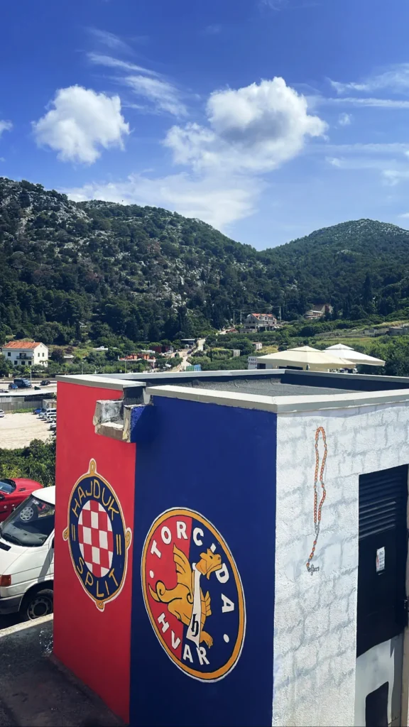 Mural de la Torcida Hvar y escudo del HNK Hajduk Split pintados en un edificio con paisaje montañoso de Dalmacia de fondo.