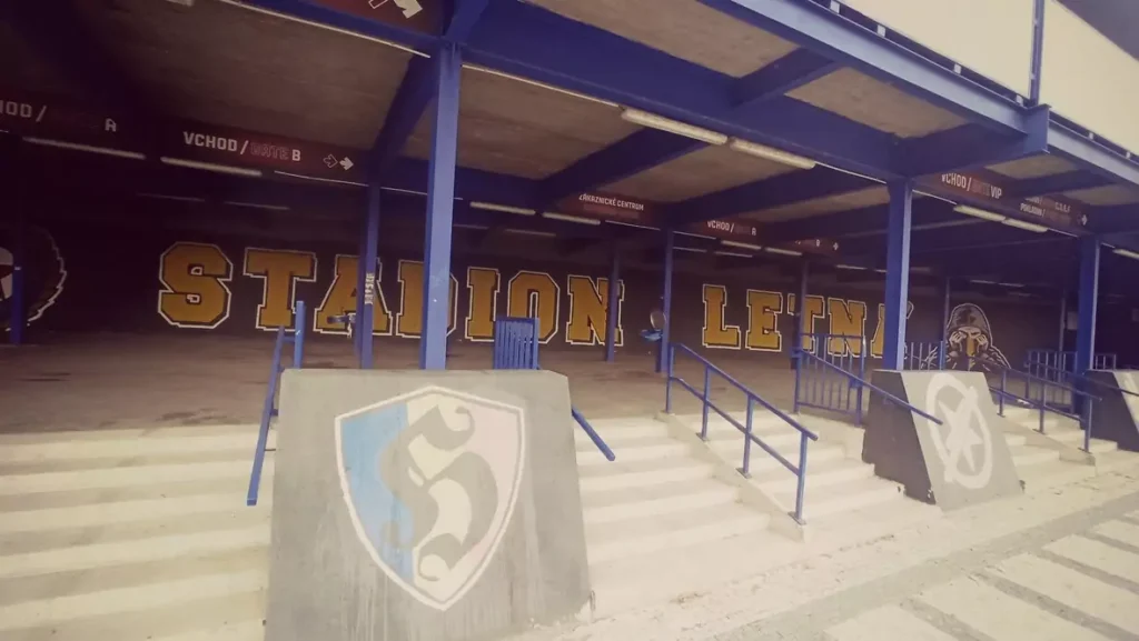 mural-hooligans-sparta-praga Vista panorámica de la entrada al estadio Letná del Sparta Praga llena de graffitis. Se lee "STADION LETNÁ" en el muro, y en primer plano hay bloques de hormigón con el escudo del Sparta y la estrella del Slavia Praga tachada.
