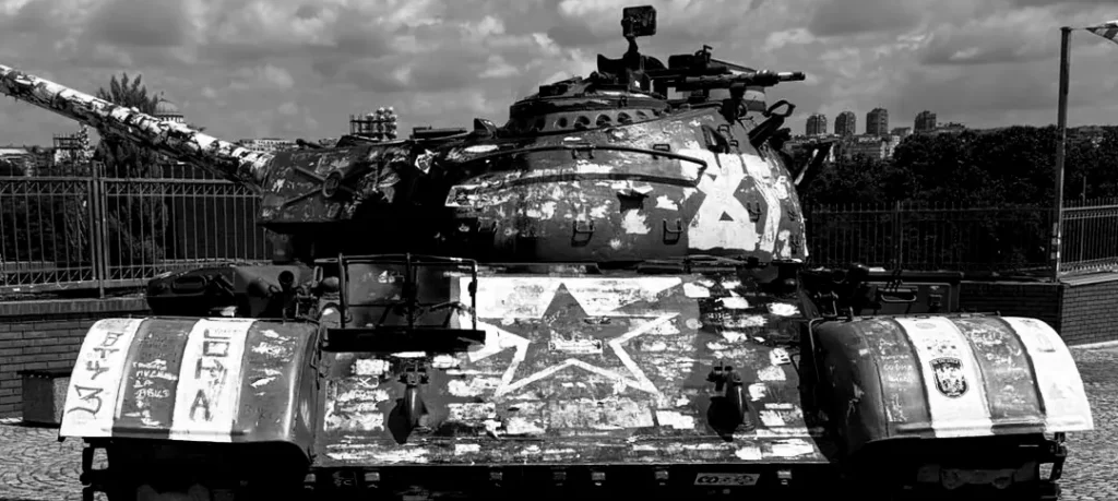 Tanque militar T-55 con camuflaje y emblemas del Estrella Roja de Belgrado (Delije) frente al estadio Rajko Mitić en blanco y negro.