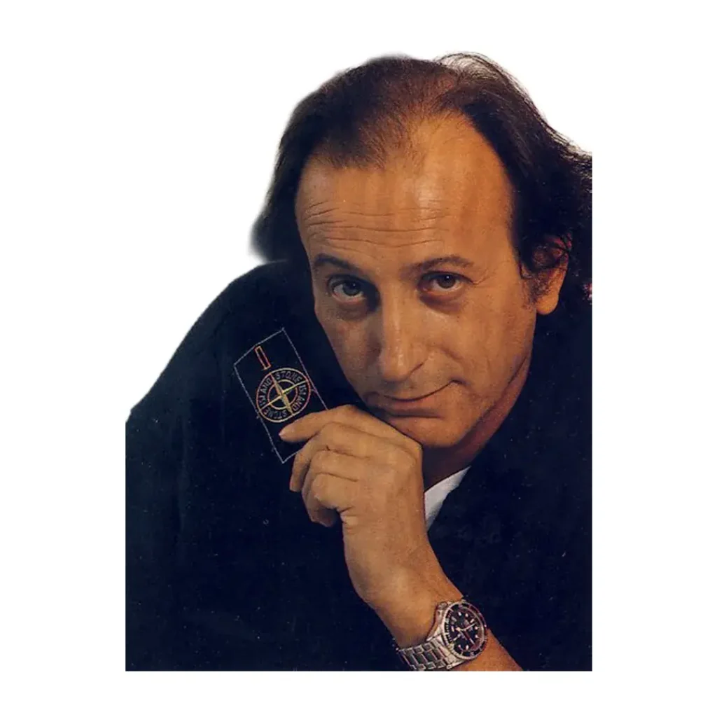 Retrato de Massimo Osti, fundador de Stone Island y C.P. Company, sujetando el icónico parche de la brújula (badge)